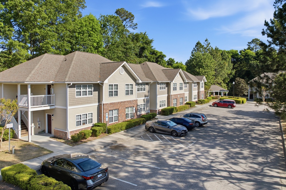 cover-photo-Fairview-Pointe-Apartments - Keystone Two-story beige apartment building with brick accents, surrounded by green trees and a paved parking lot with several cars under a blue sky